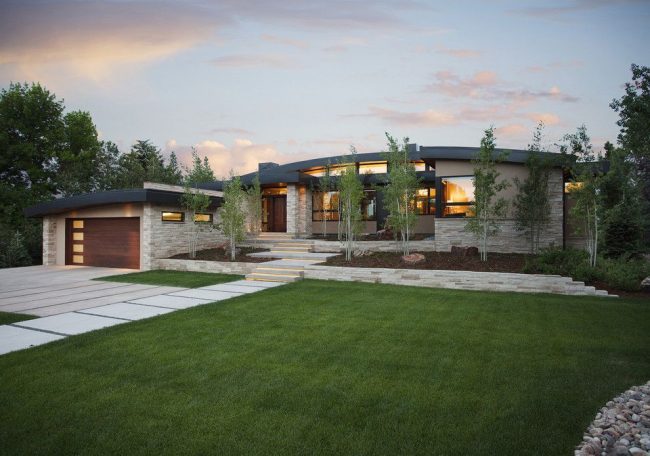 Современный экстерьер дома с гаражом: каменная облицовка, изогнутая крыша, яркое освещение, зеленая придомовая территория